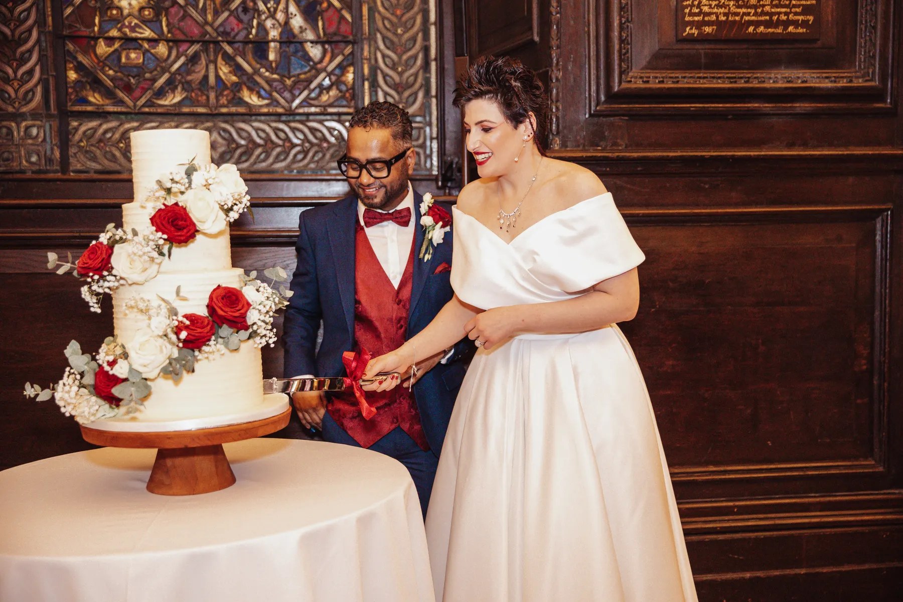 A bride in a white gown and a groom in a navy suit with a red waistcoat smile as they cut a white tiered wedding cake adorned with red and white flowers on a wooden stand at Stationers' Hall.