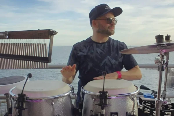 Percussionist, Andy Konga, wearing sunglasses and a cap plays percussion instruments outdoors on a stage by the water, with a scenic sea view in the background under a partly cloudy sky.