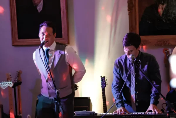 from Indie/Britpop covers band, Indie Five, perform indoors under bright lights, with a singer at the mic, a keyboard player, and guitarists. Portrait paintings hang on the wall behind them.