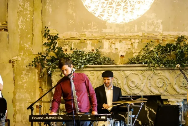 Indie/Britpop covers band, Indie Five, performs indoors; one plays keyboard and sings, another plays drums, while two others play guitar and bass. The backdrop is an ornate, weathered wall with greenery and a large, glowing light overhead.