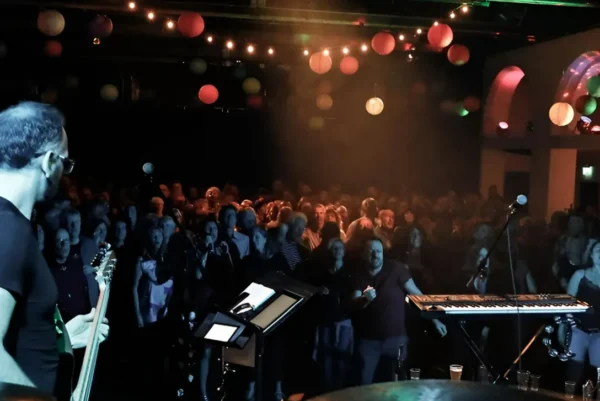 A musician from Indie/Britpop covers band, Indie Five, with a guitar stands on stage, facing a large, cheering crowd in a dimly lit venue decorated with colourful hanging lights and paper lanterns. Musical instruments and equipment are visible onstage.