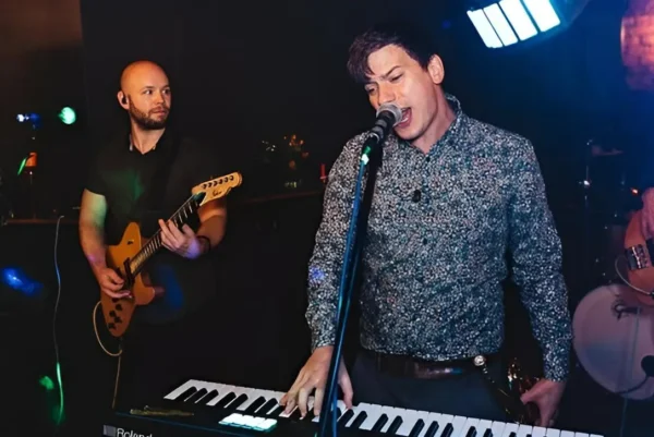 Indie/Britpop covers band, Indie Five, performs on stage; a man sings into a microphone while playing a keyboard, accompanied by a guitarist and bassist under colourful stage lights in a dimly lit venue.