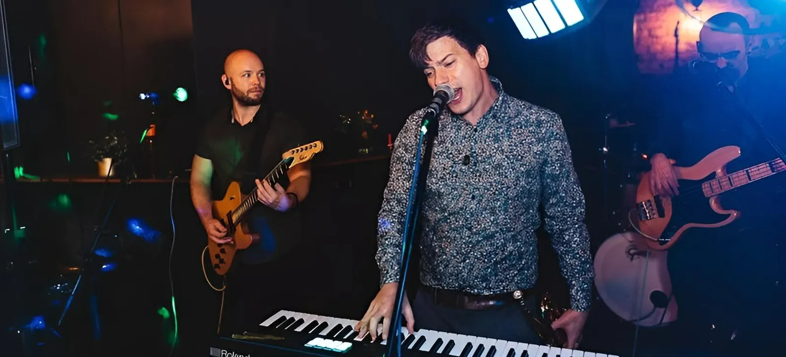 Indie/Britpop covers band, Indie Five, performs on stage; a man sings into a microphone while playing a keyboard, accompanied by a guitarist and bassist under colourful stage lights in a dimly lit venue.