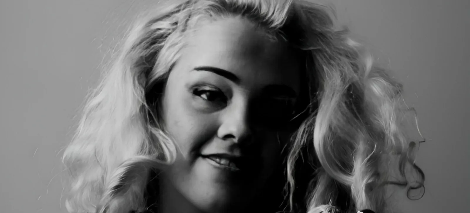 Black and white close-up portrait of a person with wavy blonde hair, slightly smiling, looking towards the camera, with soft, dramatic lighting creating shadows on their face.