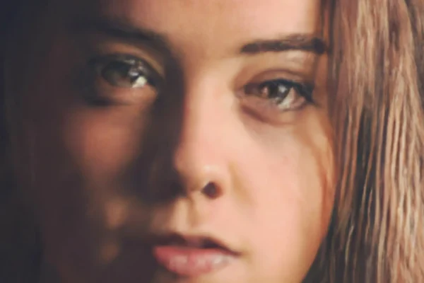 A young woman with long brown hair and light skin looks directly at the camera with a neutral expression. The lighting is soft, highlighting her facial features and eyes.