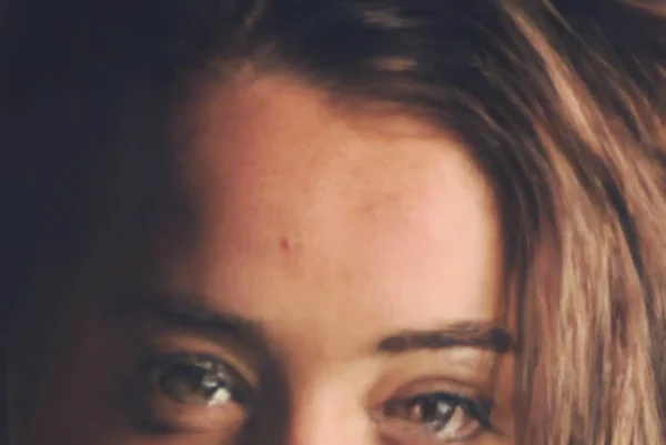 A young woman with long brown hair and light skin looks directly at the camera with a neutral expression. The lighting is soft, highlighting her facial features and eyes.