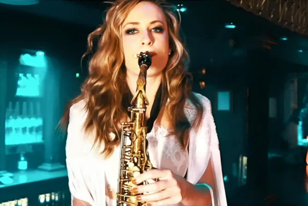 A woman with long wavy hair plays a saxophone in a dimly lit, blue-toned indoor setting, wearing a white top and looking directly at the camera.