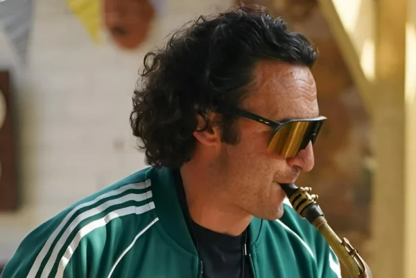 A man with curly hair and sunglasses wearing a green Adidas jacket plays a saxophone indoors.