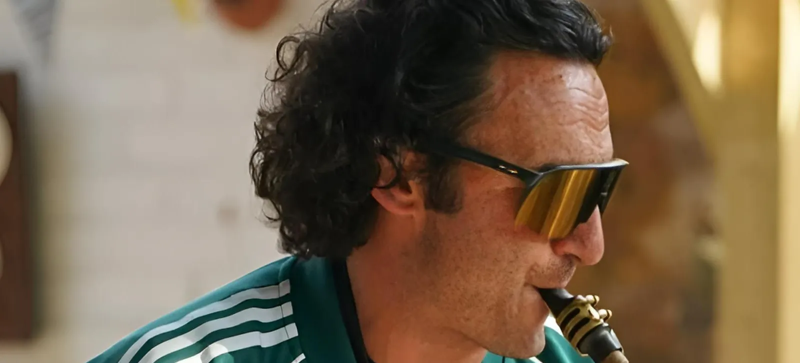 A man with curly dark hair and sunglasses plays a wind instrument. He is wearing a green and white striped shirt and is outdoors, with a blurred background.