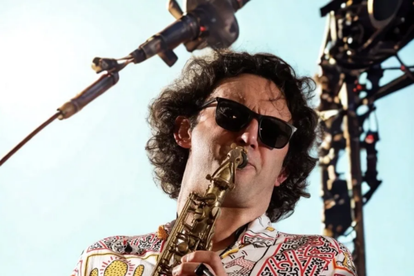 A man wearing sunglasses and a colourful patterned shirt plays a saxophone on stage, with stage lights and equipment visible in the background.