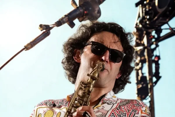 A man wearing sunglasses and a colourful patterned shirt plays a saxophone on stage, with stage lights and equipment visible in the background.