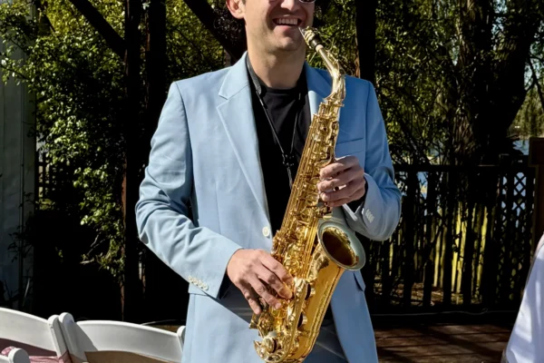 A man in a light blue suit and sunglasses smiles whilst playing a gold saxophone outdoors near white chairs and greenery.