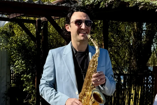 A man in a light blue suit and sunglasses smiles whilst playing a gold saxophone outdoors near white chairs and greenery.