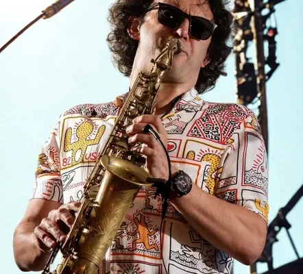 A musician wearing sunglasses and a colourful patterned shirt plays a saxophone on stage, with bright lighting and concert equipment visible in the background.