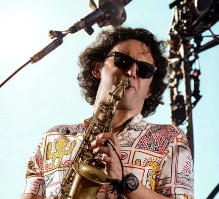A musician wearing sunglasses and a colourful patterned shirt plays a saxophone on stage, with bright lighting and concert equipment visible in the background.