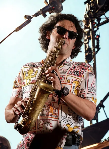 A musician wearing sunglasses and a colourful patterned shirt plays a saxophone on stage, with bright lighting and concert equipment visible in the background.