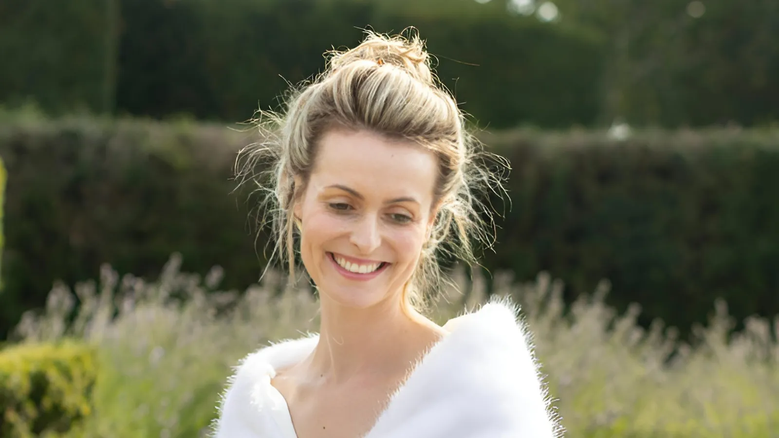 A smiling woman with blonde hair in a loose updo stands outdoors, wearing a white, fur-trimmed shawl. Green foliage and lavender plants are blurred in the background.