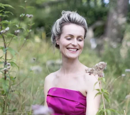 A woman in a strapless, bright pink dress stands smiling among tall grass and wildflowers in an outdoor setting. She has fair hair styled up and looks down at a plant, appearing relaxed and happy.