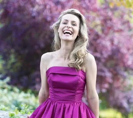 A woman with long blonde hair wearing a strapless, bright pink dress stands outdoors, smiling and laughing by a stone wall, with lush greenery and purple-leaved trees in the background.
