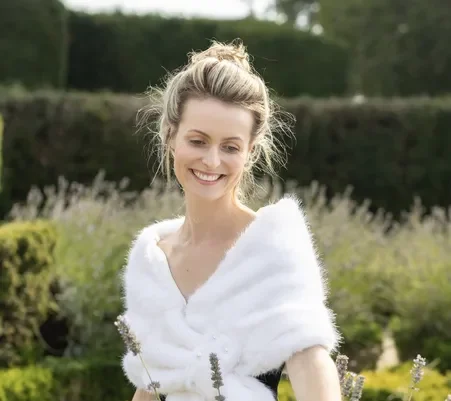 Classical soprano singer, Millicent, blonde hair in an updo, wearing a white fur stole and dark dress, stands in a garden and gently touches lavender flowers, smiling. Boxwood hedges and greenery are in the background.