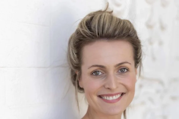 A woman with blonde hair in an updo, wearing a strapless black dress, smiles whilst leaning against a white wall with decorative moulding.