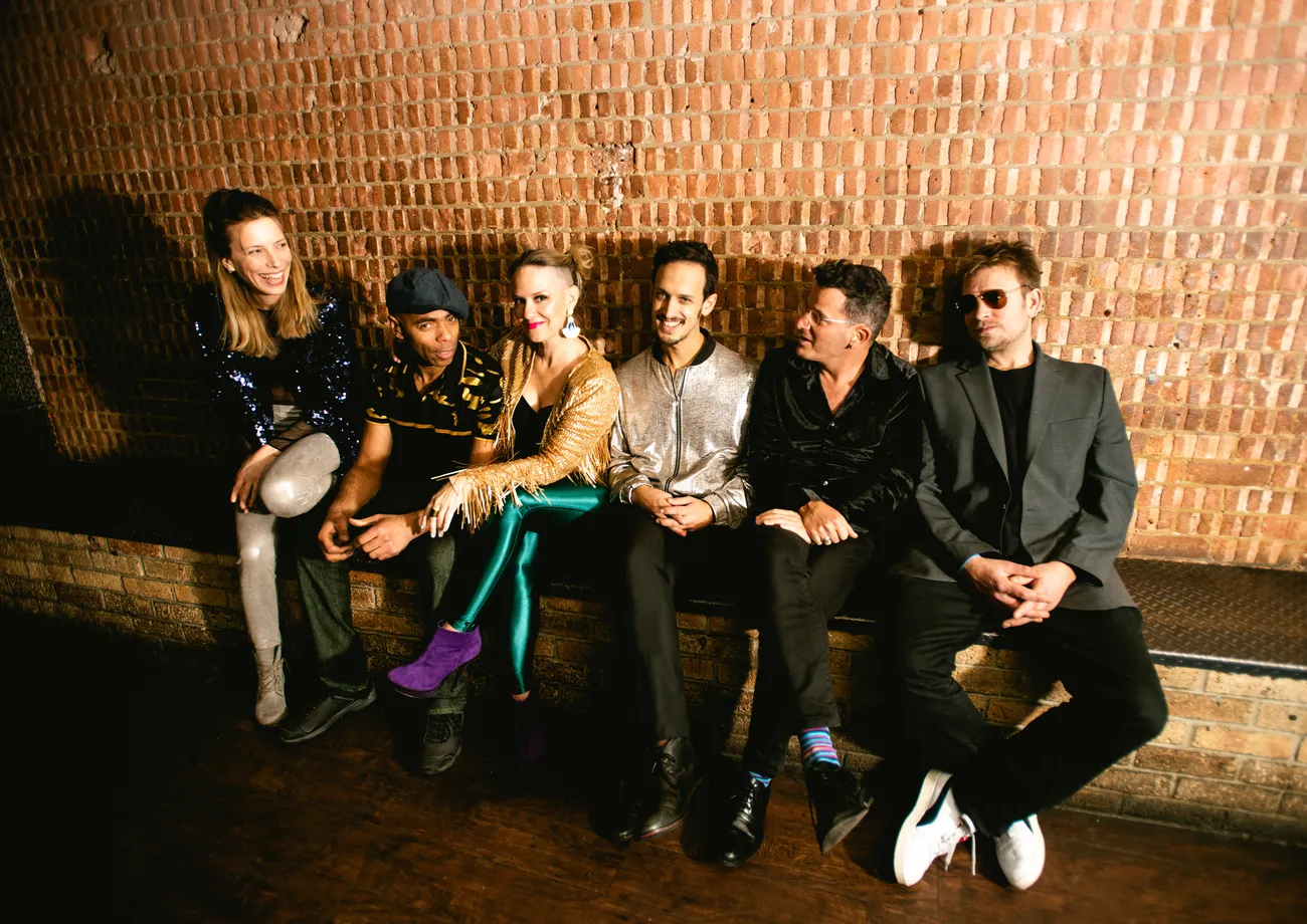Six people sit on a bench against a brick wall, dressed in colourful, eclectic outfits, appearing joyful and engaged in conversation, with some smiling and laughing.