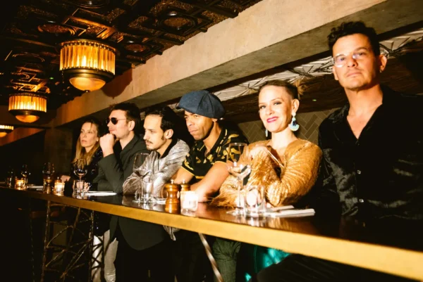 Six people sit side by side at a bar counter in a warmly lit, stylish venue, looking towards the camera. They wear fashionable, eclectic outfits and appear relaxed and engaged.