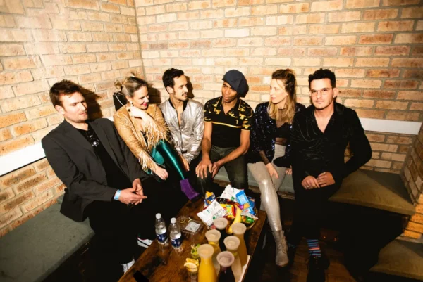 Six people sit on benches in a cosy, brick-walled room, chatting and smiling. A table in front holds snacks, drinks, and water bottles. The group appears relaxed and casual, dressed in colourful, stylish outfits.