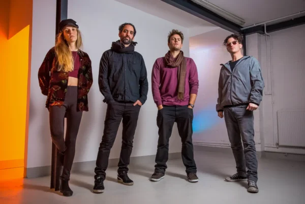 Four people stand indoors against a plain wall, posing confidently. Two men and one woman wear casual, dark clothing, and another man wears a grey jacket and sunglasses. The lighting has orange and blue accents.
