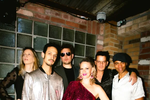 Six people pose together indoors against a brick and glass wall, wearing stylish, shiny party outfits. One woman stands in front in a sparkly red dress, whilst the others wear metallic or patterned clothing and smile at the camera.