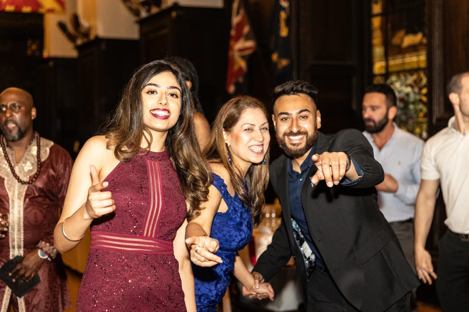 A group of people dressed formally smile and pose for the camera at an indoor event or party, with three people in front pointing and appearing cheerful. An event DJ entertains as other guests mingle in the background.
